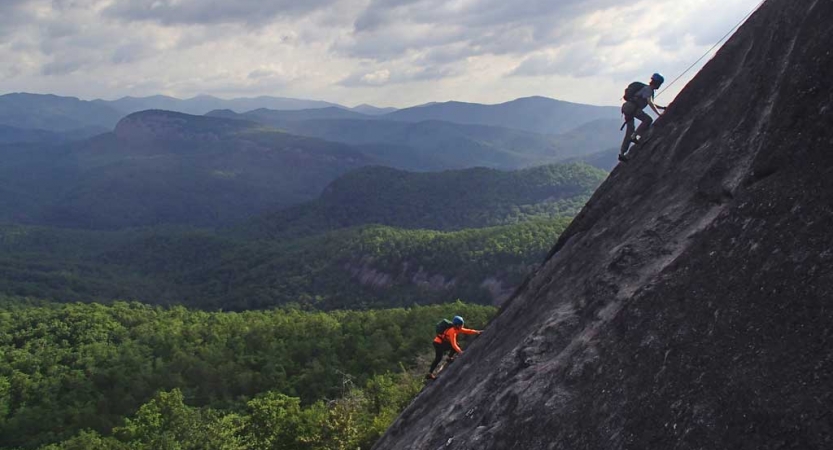 Two people wearing safety gear are secured by ropes as they each climb a steep rock face. The blue ridge mountains are below them. 